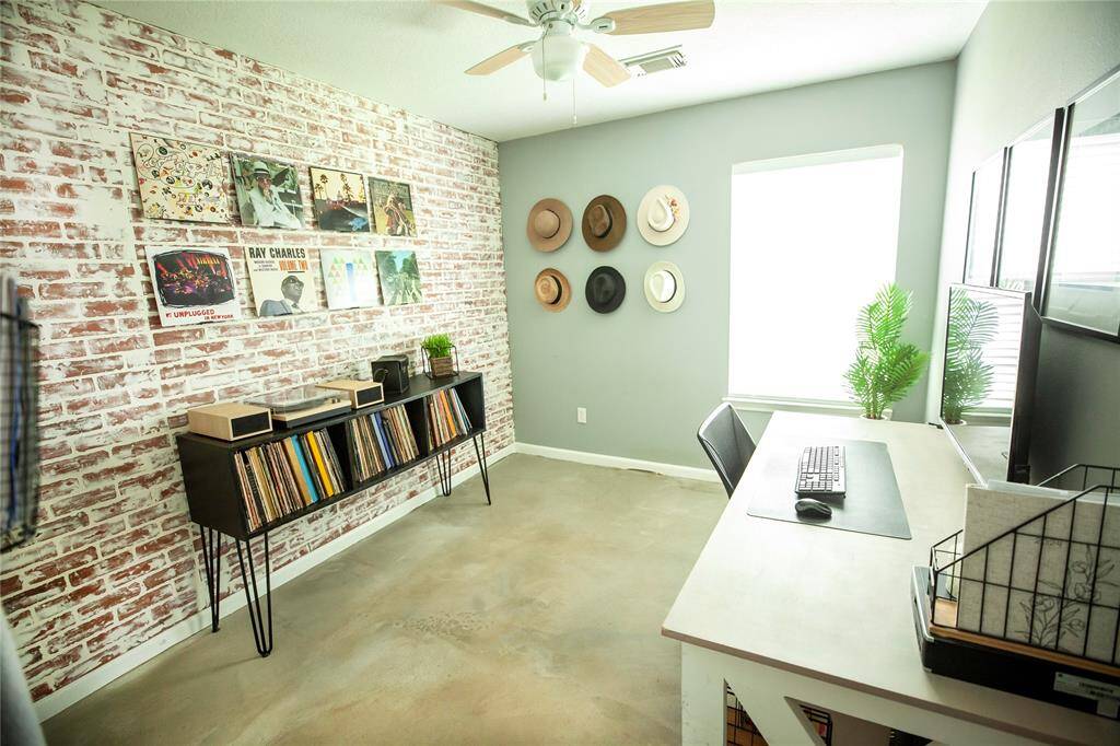 This is one of the bedrooms that have been transformed into a unique home office with a modern industrial vibe, showcasing a faux brick accent wall, vinyl record display, and ample natural light.