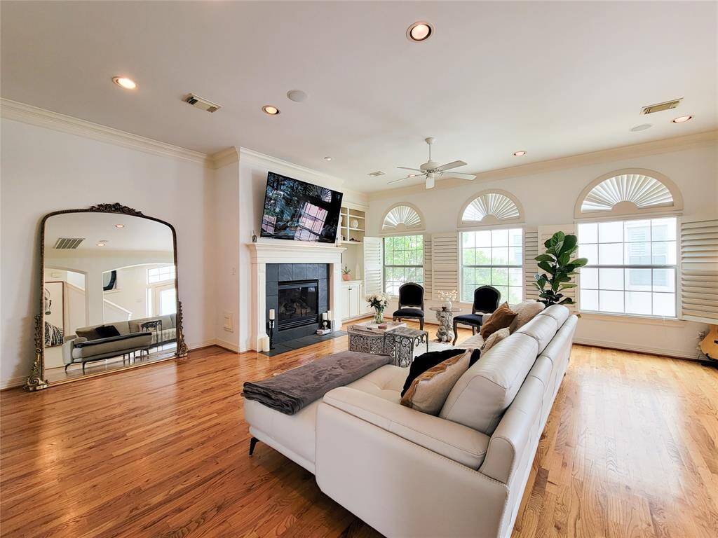 Bright and spacious living room with gleaming hardwood floors, featuring a cozy fireplace and a mounted TV. Large windows bring in natural light, and elegant arched shutters add charm.