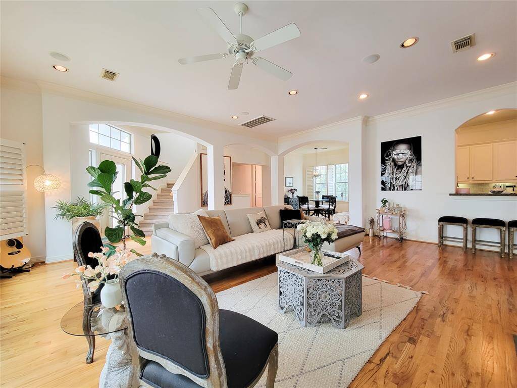This bright and airy living space features hardwood floors, high ceilings, recessed lights stylish decor, and ample natural light. An open floor plan connects the living room to a dining area and kitchen, while large archways and a staircase add architectural interest. Perfect for comfortable living and entertaining.