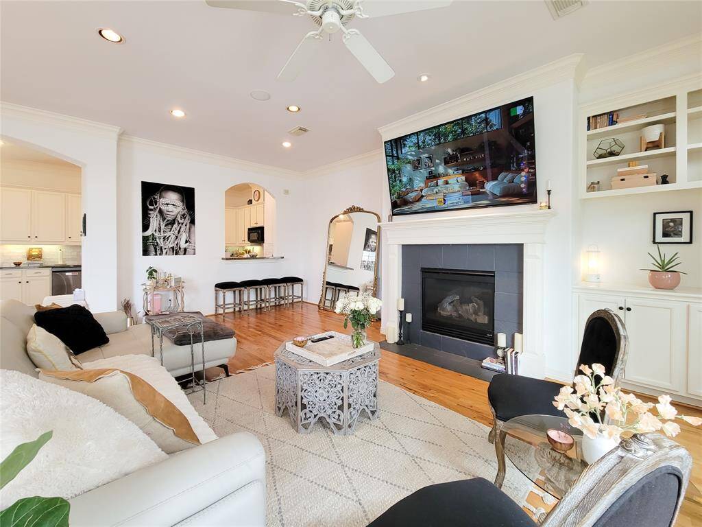 Bright and inviting living room featuring hardwood floors, a cozy fireplace, and built-in shelves. The open layout connects to the kitchen, and a large TV is mounted above the fireplace. Elegant decor and ample natural light create a welcoming atmosphere.