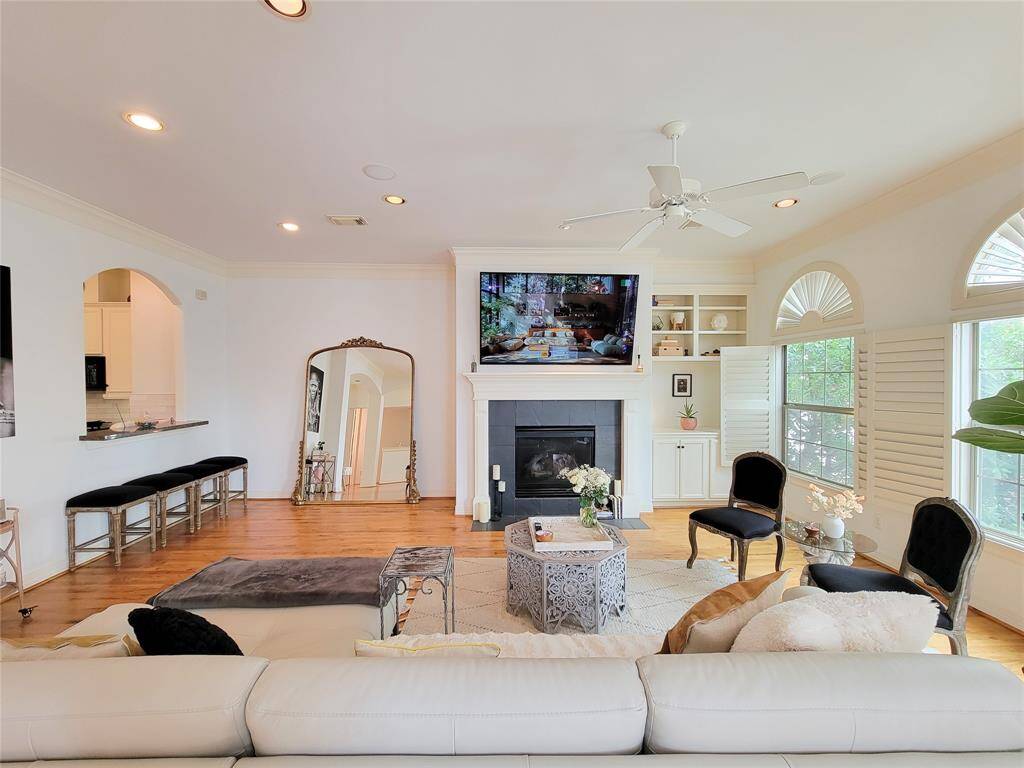 Bright and inviting living room with hardwood floors, a central fireplace, built-in shelves, and large windows with shutters. The open layout includes a pass-through to the kitchen with bar seating, and a ceiling fan adds comfort. Perfect for relaxation and entertaining.
