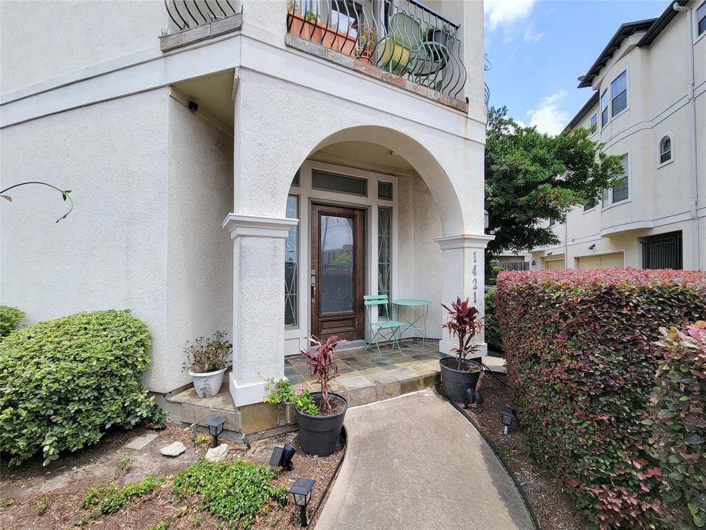 This photo shows a welcoming entrance to a townhouse with an arched doorway, small patio area with seating, and neatly landscaped bushes.