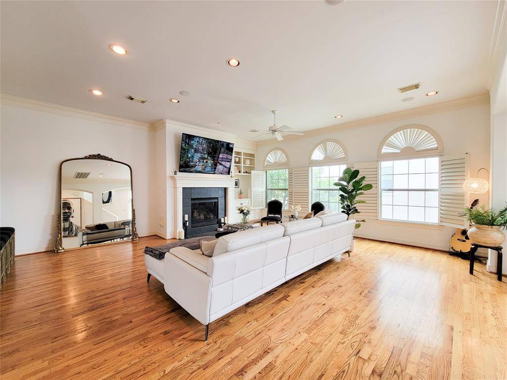 This spacious living room features hardwood floors, a cozy fireplace, and large windows with shutters providing ample natural light. It includes elegant furniture, a large mirror, and tasteful decor, creating a warm and inviting atmosphere.