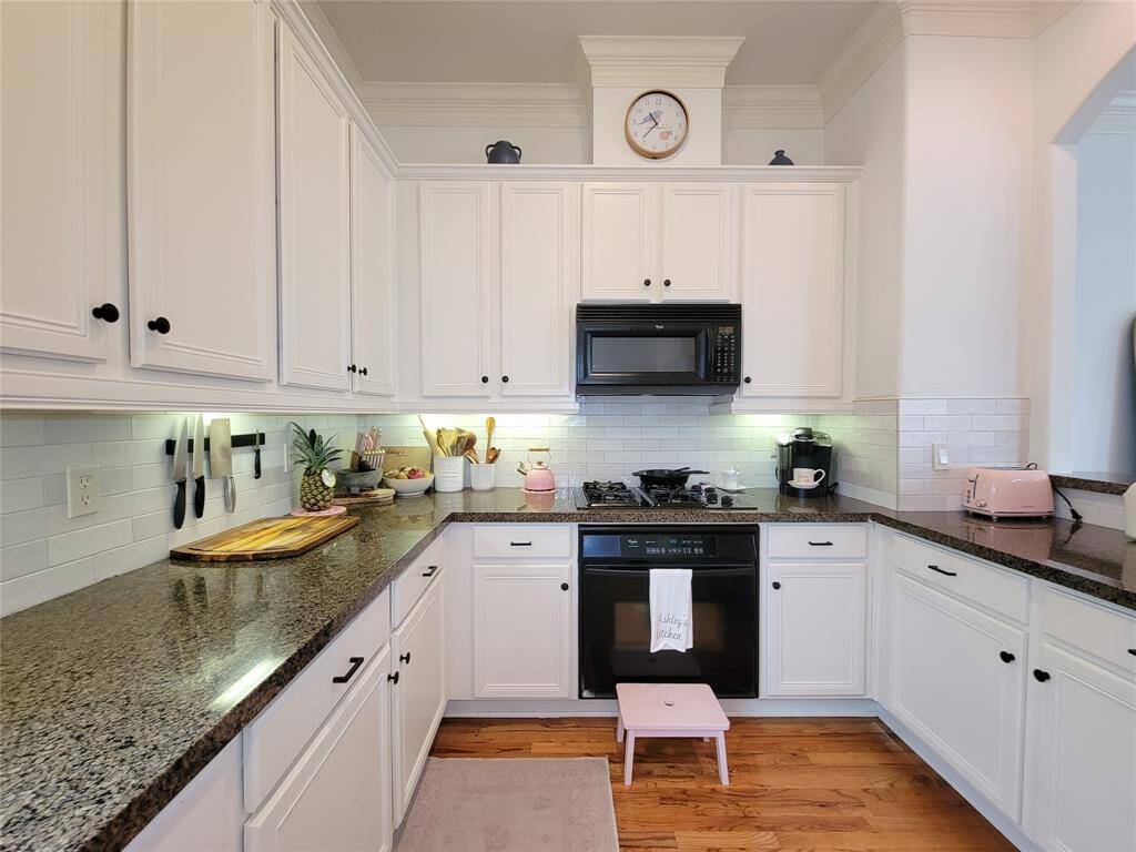 This kitchen features white cabinetry with dark granite countertops and a subway tile backsplash. It includes a black oven and microwave, a gas cooktop, and ample storage. The space is bright with under-cabinet lighting and has wood flooring, and pass through open to living room.