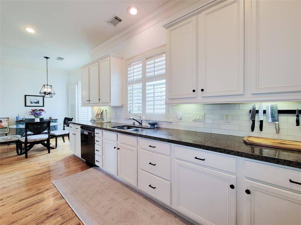 This bright kitchen features white cabinetry, granite countertops, and a subway tile backsplash. It includes a dining area with a modern chandelier, hardwood floors, and ample natural light through large windows.