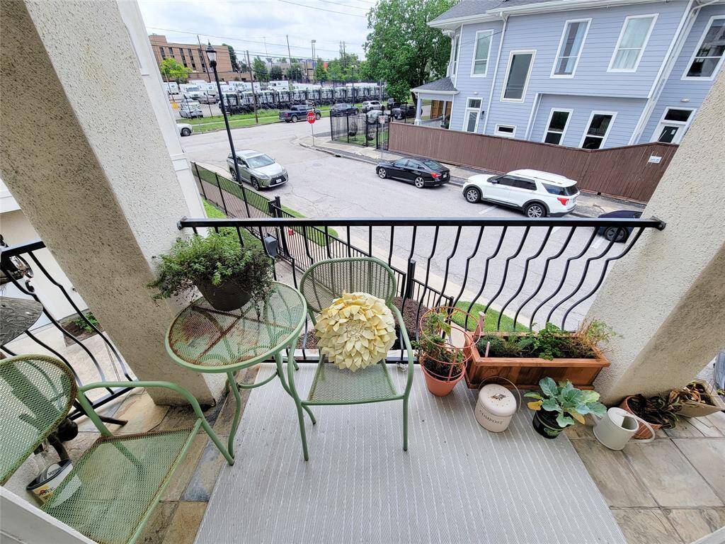 Cozy balcony with a small table and chairs, surrounded by potted plants. Overlooks a quiet street with a view of neighboring buildings and parked cars. Ideal for relaxing or enjoying a morning coffee.