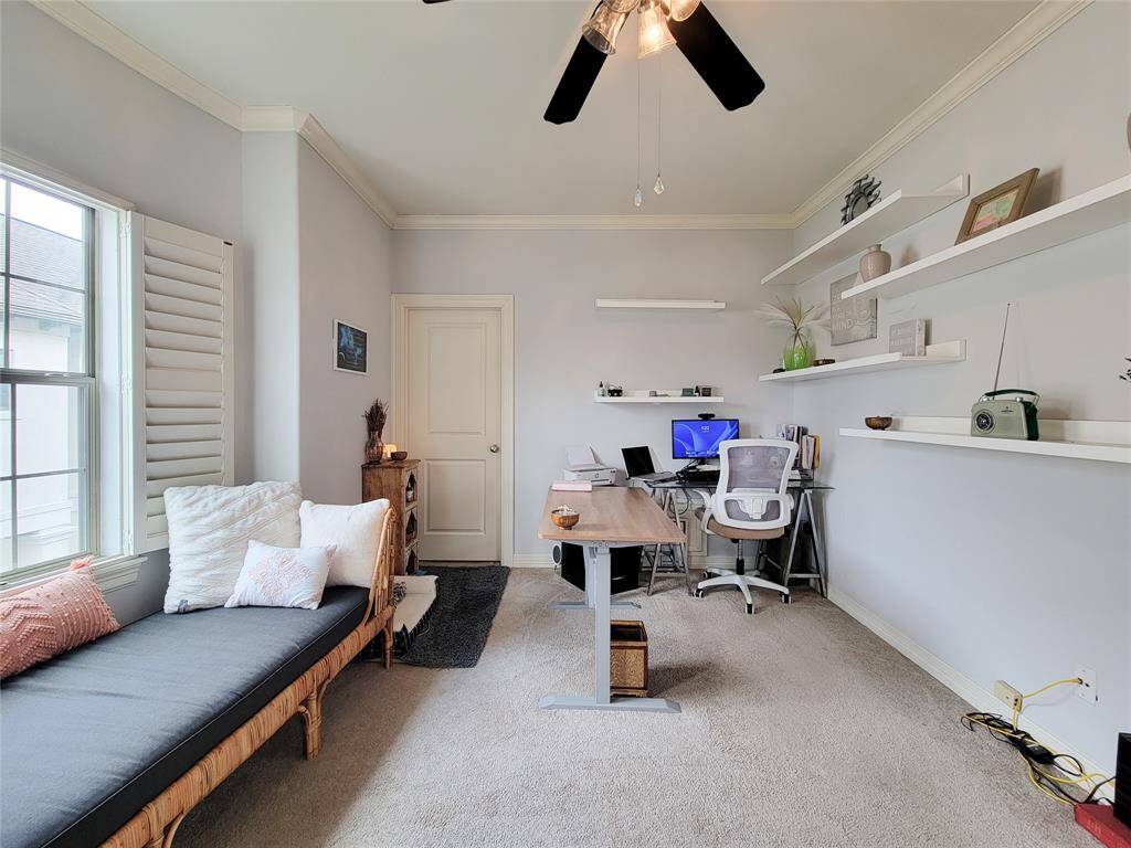 This room is a bright, well-lit office space with a large window and plantation shutters. It features a neutral color scheme, a modern desk setup, floating shelves for storage, and a comfortable seating area. Ideal for a productive work-from-home environment.
