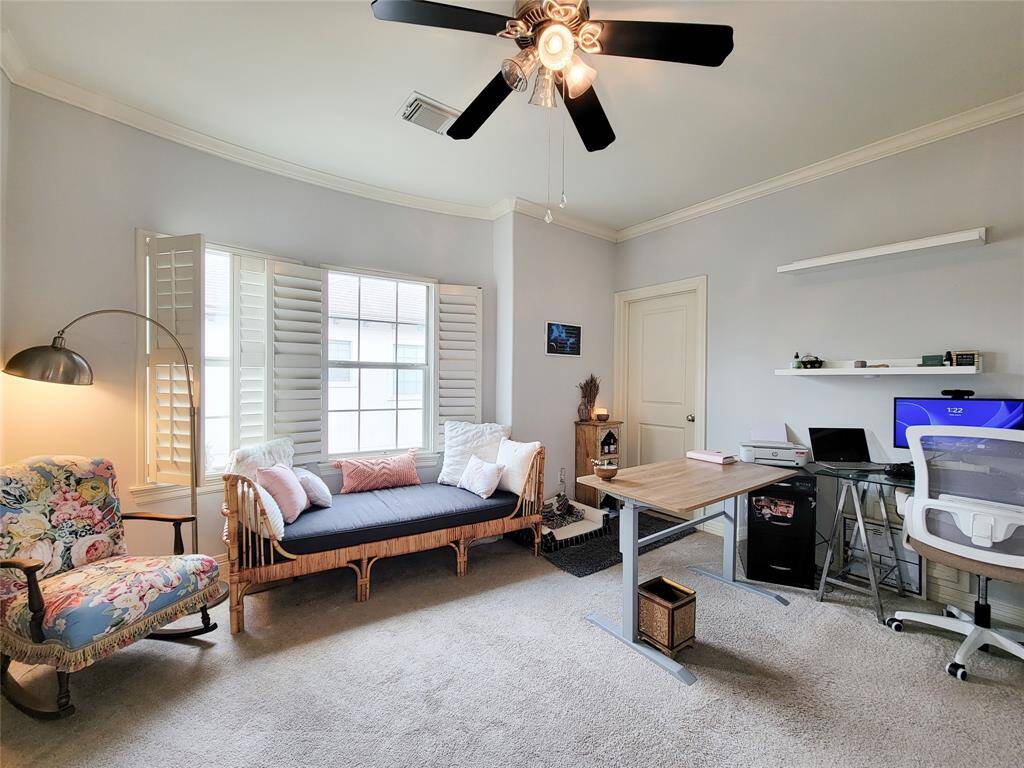 Bright home office with large windows and shutters, featuring a cozy daybed, modern desk, and comfortable seating. Neutral walls and carpeting create a calming atmosphere, while a ceiling fan provides additional comfort.