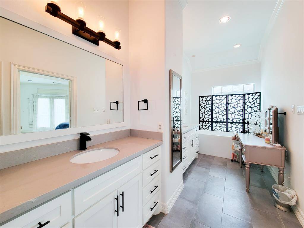 This bright bathroom features a large mirror with modern lighting, a spacious vanity with ample storage, sleek countertops, and decorative privacy screens near the window, creating a stylish and functional space.