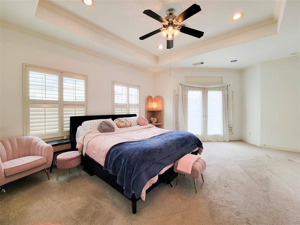 Spacious, bright bedroom with a tray ceiling, featuring a large bed, plush carpeting, and elegant pink accent chairs. Ample natural light from windows with shutters and a glass door.