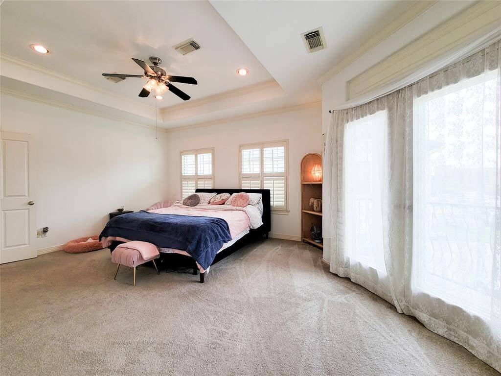 This spacious bedroom features a cozy bed with elegant bedding, a ceiling fan, and ample natural light from large windows with shutters. The room has soft carpeting and a door leading to a private balcony, offering a peaceful retreat.