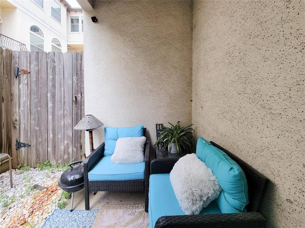 Cozy outdoor patio with privacy fencing, featuring comfortable wicker seating with blue cushions, a small grill, and decorative plants. Ideal for relaxing or entertaining.