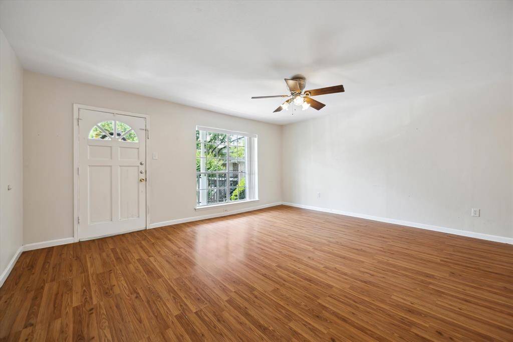 Another view of your bright, spacious living room with gorgeous floors and large window for natural light. This unit features a welcoming front door and neutral walls, lush greenery outside with the perfect balance of sun and shade.