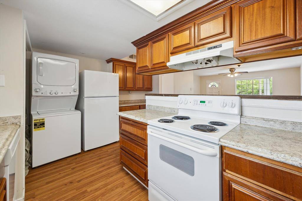 This kitchen features warm wooden cabinetry, a white electric stove, and stacked washer and dryer units. It has ample countertop space and a cozy, open layout that extends into a bright living area.