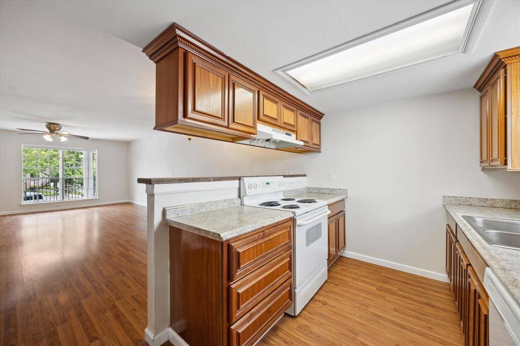 This photo showcases a bright, open-concept kitchen and living area with beaming floors. The kitchen features ample counter space and plenty of storage! The living area has a ceiling fan and large windows overlooking the courtyard below providing a welcoming and airy feel.