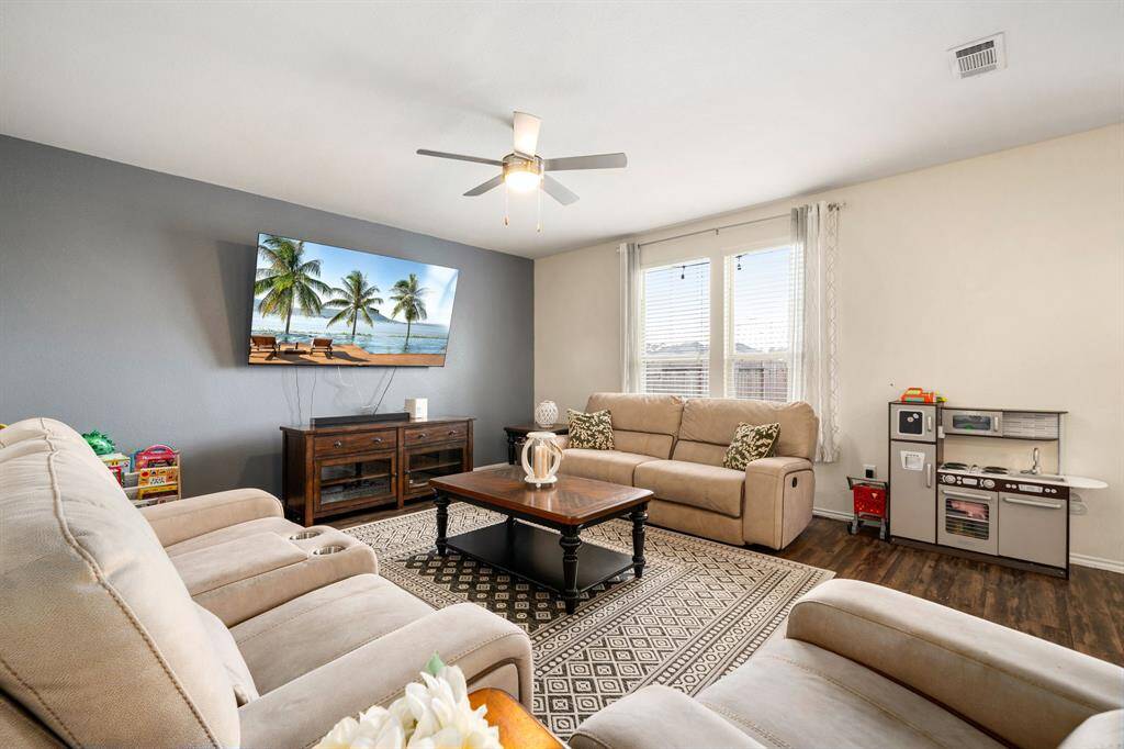 Spacious living room with wood-style flooring, a ceiling fan, and large windows that fill the space with natural light. The neutral tones and accent wall create a warm, comfortable setting perfect for everyday living and entertaining.