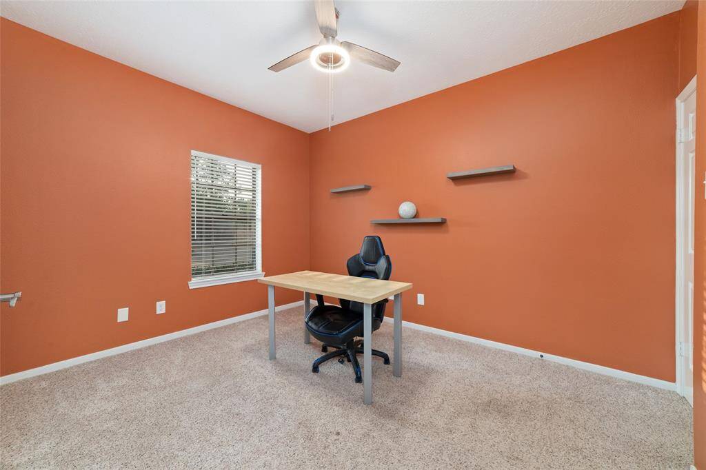 Bright home office with vibrant orange walls, a window with blinds, and a ceiling fan. Features a minimalist desk, office chair, and wall-mounted shelves. Cozy carpeted flooring enhances the inviting atmosphere.