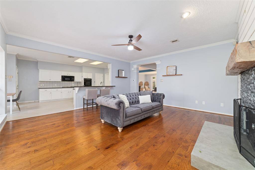 This spacious living area features hardwood floors, a cozy fireplace with a rustic wooden mantle, and an open concept leading into a modern kitchen with white cabinetry and a breakfast bar.