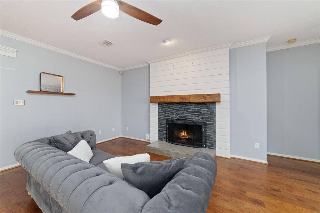 Cozy living room with hardwood floors, a stylish gray sectional sofa, and a modern fireplace featuring a white shiplap surround and wooden mantel. Soft blue walls and a ceiling fan add to the inviting atmosphere.