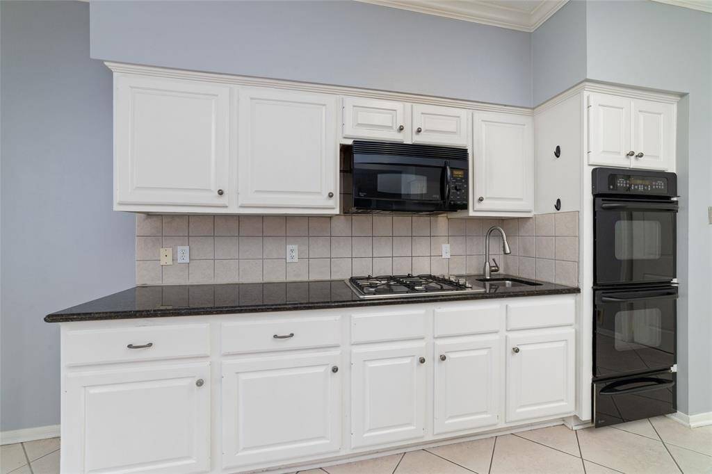 This kitchen features white cabinetry, a sleek black countertop, and a tiled backsplash. It includes a built-in gas cooktop, black microwave, and double oven, with a modern sink and faucet for convenience.