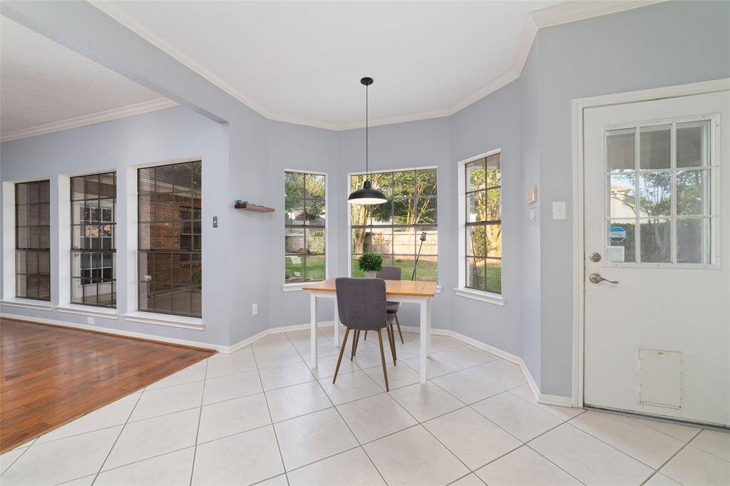 This bright space features a cozy breakfast nook with large windows overlooking the backyard. The room has a modern, open layout with light gray walls, tile flooring, and a transition to hardwood, offering a welcoming atmosphere.
