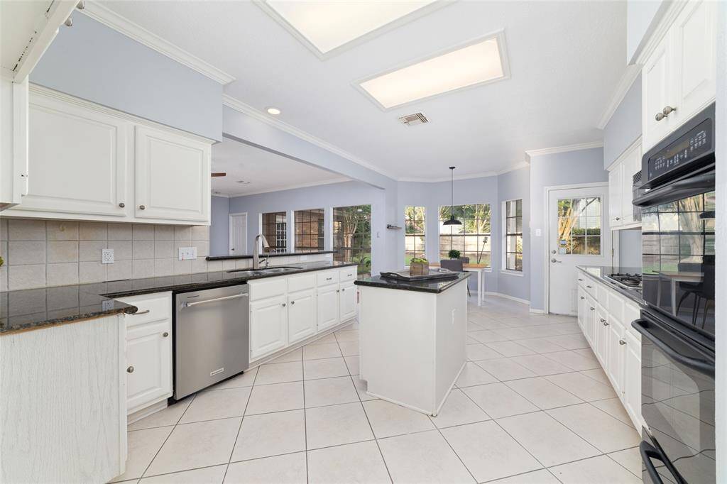 Bright, spacious kitchen with white cabinetry, black countertops, and a central island. Features include modern appliances, tile flooring, and large windows that offer natural light and garden views. An adjoining breakfast nook enhances the open layout.