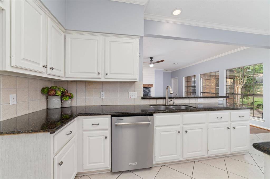 This bright kitchen features white cabinets, dark granite countertops, and a modern stainless steel dishwasher. It opens to a spacious living area with large windows offering plenty of natural light and a view of the backyard.