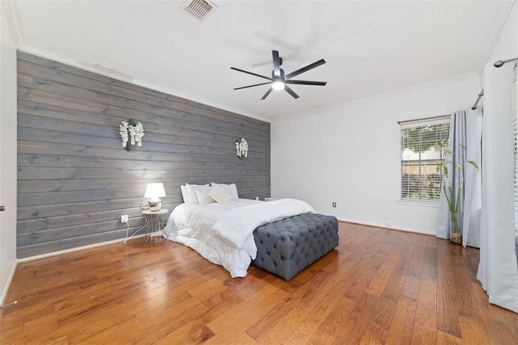 This spacious bedroom features elegant wood flooring, a stylish gray accent wall, and ample natural light from a large window. The modern ceiling fan and cozy bedding create a relaxing atmosphere.