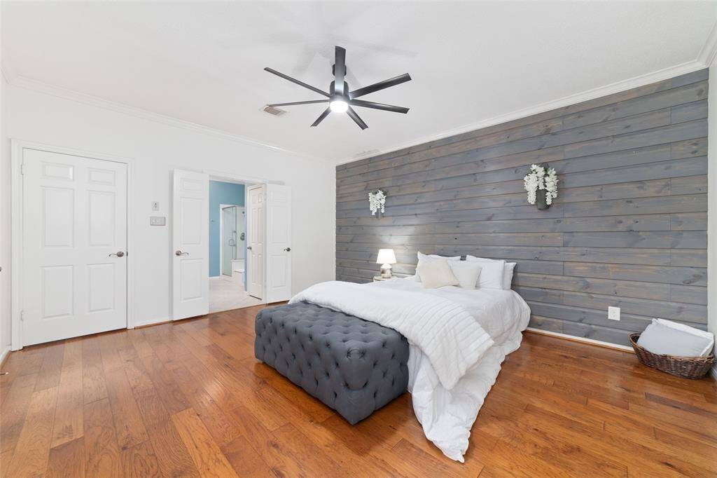 This spacious bedroom features a stylish wood accent wall, warm hardwood floors, and modern ceiling fan. It includes a cozy bed setup with a plush bench and opens to a bathroom with double doors, creating a serene and inviting space.