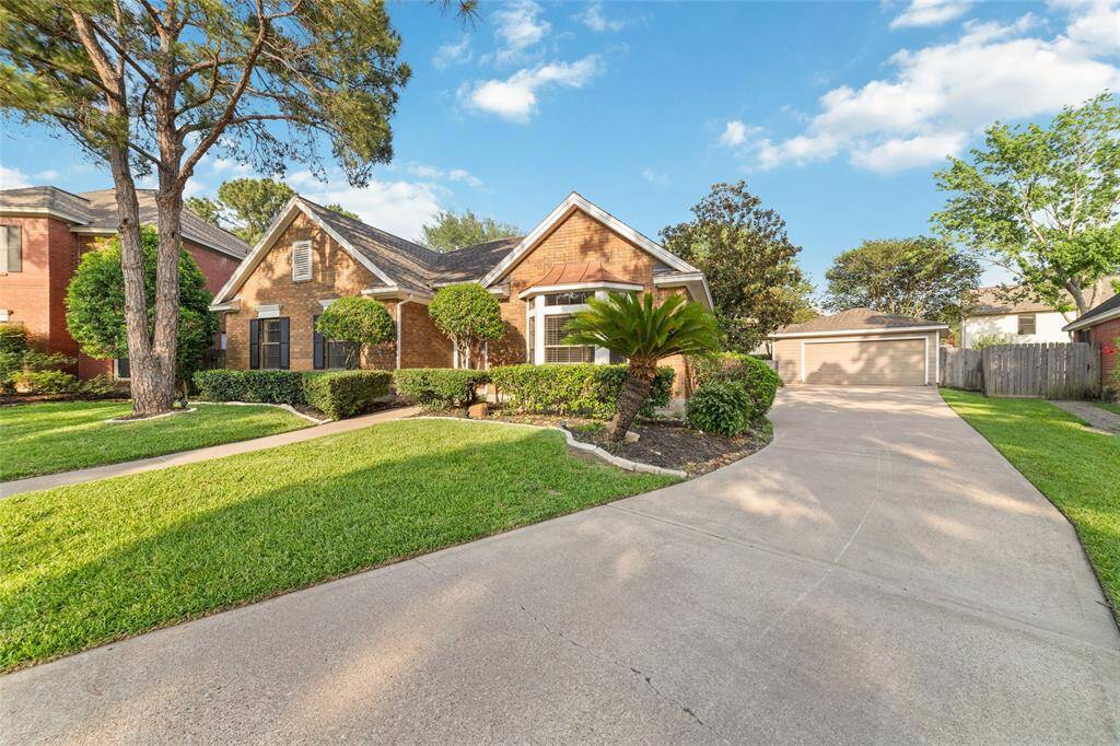 Charming brick home with a manicured front yard, featuring lush greenery and a wide driveway leading to a detached garage. The property offers a welcoming curb appeal in a serene neighborhood setting.