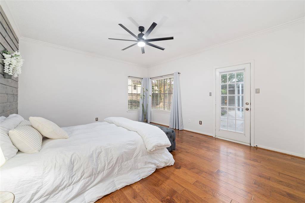 This bright, airy bedroom features hardwood floors, a stylish ceiling fan, and a cozy bed with neutral bedding. Large windows and a glass door provide plenty of natural light and access to the outdoor space.