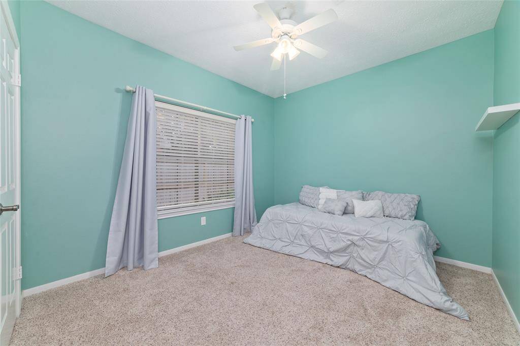 This room features soft teal walls, a cozy carpeted floor, and a ceiling fan. It includes a window with light curtains and a daybed, creating a comfortable, inviting space.