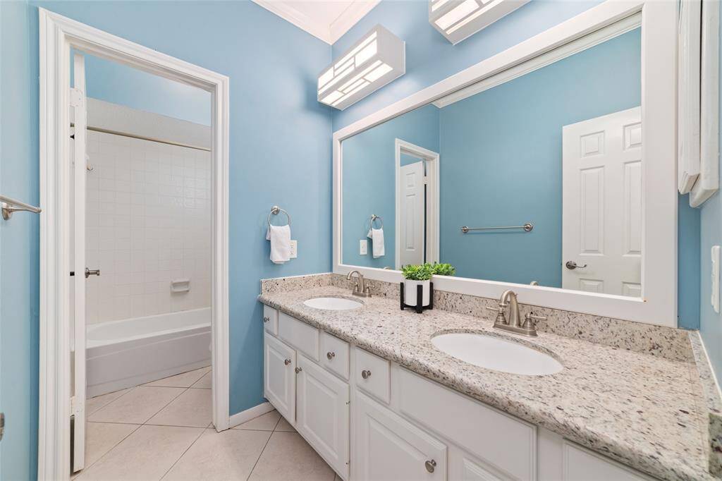 This bathroom features a double vanity with granite countertops, blue walls, and a large mirror. It has modern lighting and a bathtub with a shower in a separate tiled area.
