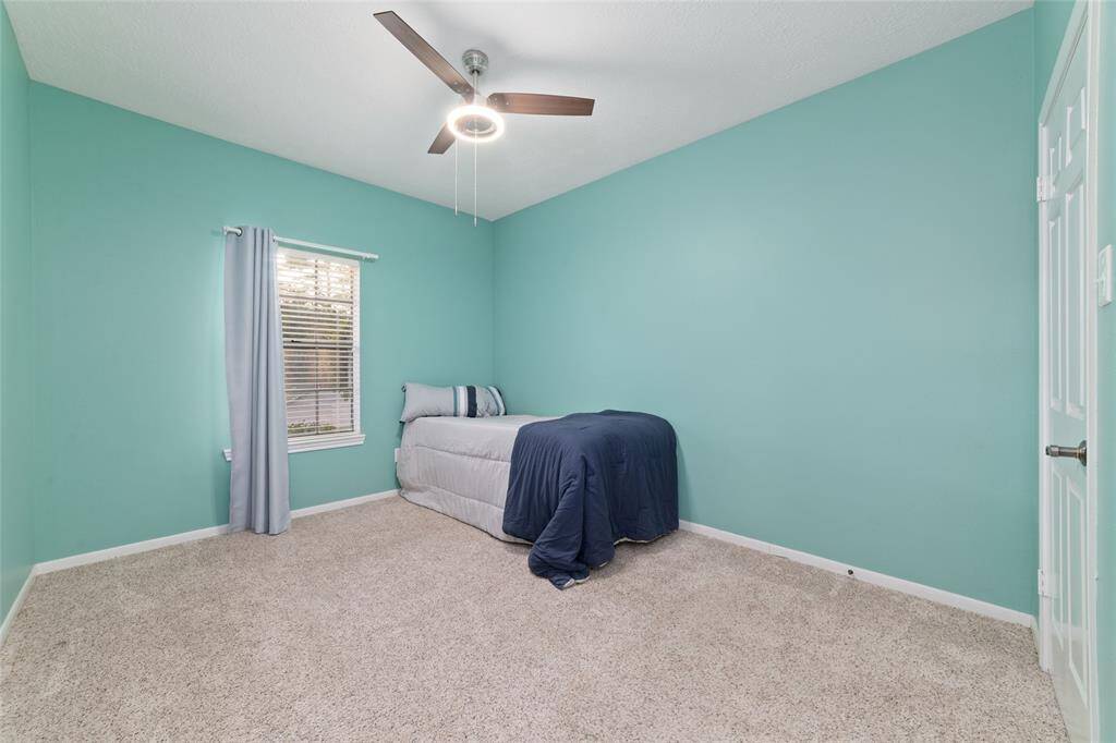 This bedroom features calming teal walls, a cozy carpeted floor, and a ceiling fan with a light. A window with light curtains provides natural light, and there's ample space for furniture.