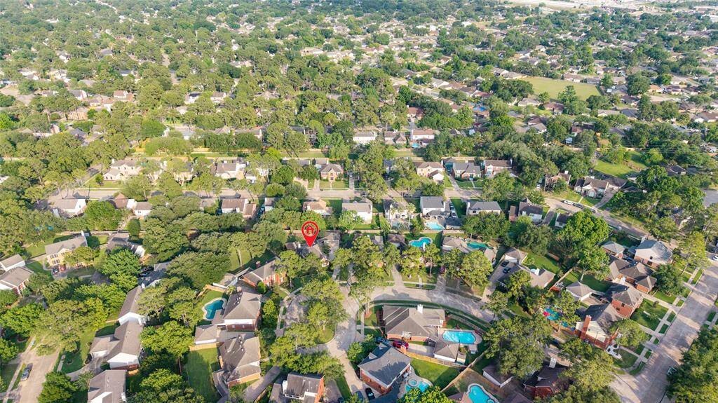 This aerial photo showcases a lush, green suburban neighborhood, featuring tree-lined streets and numerous houses with spacious yards. Many homes have swimming pools, indicating a family-friendly area with ample recreational opportunities. The neighborhood appears well-maintained and peaceful, ideal for a serene living environment.
