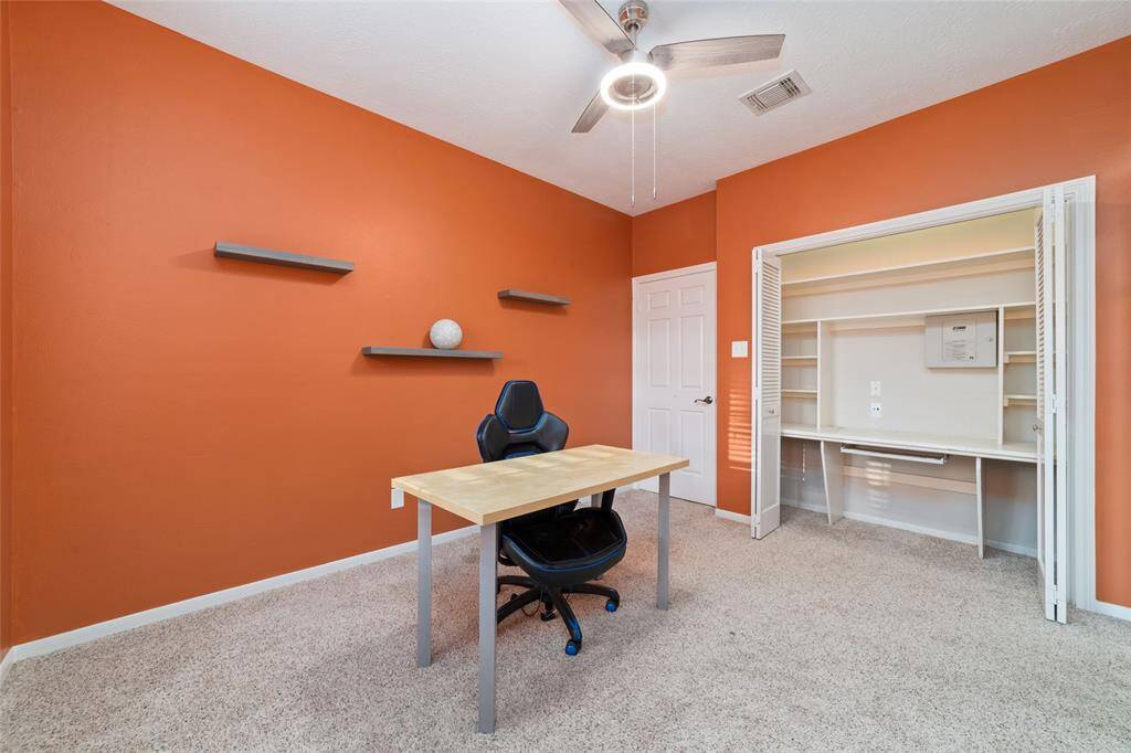 This room features a warm orange accent wall, a ceiling fan with light, and a compact work desk with a chair. It includes wall shelves for storage and a closet with built-in shelving, ideal for a home office.