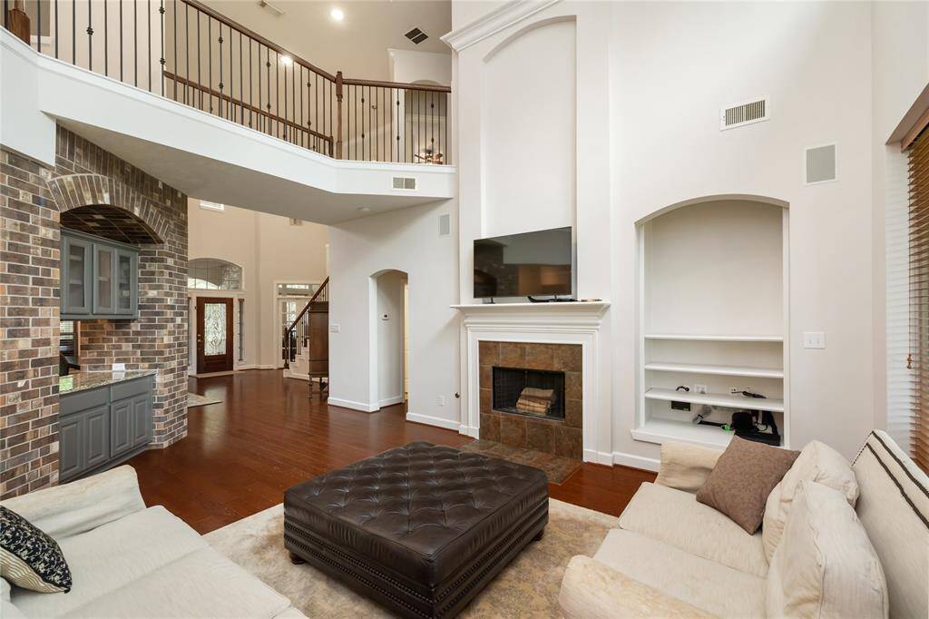 This alternate view of the spacious living room highlights the soaring two-story ceiling, elegant wrought iron staircase, and open sightlines to the front entry, private study, and serene primary suite. The cozy fireplace is flanked by built-in shelving, while the brick-accented alcove with cabinetry and granite countertop adds warmth and functional storage. Upstairs offers three additional bedrooms, a game room, and a versatile flex space that could serve as a fifth bedroom, home office, or media room.