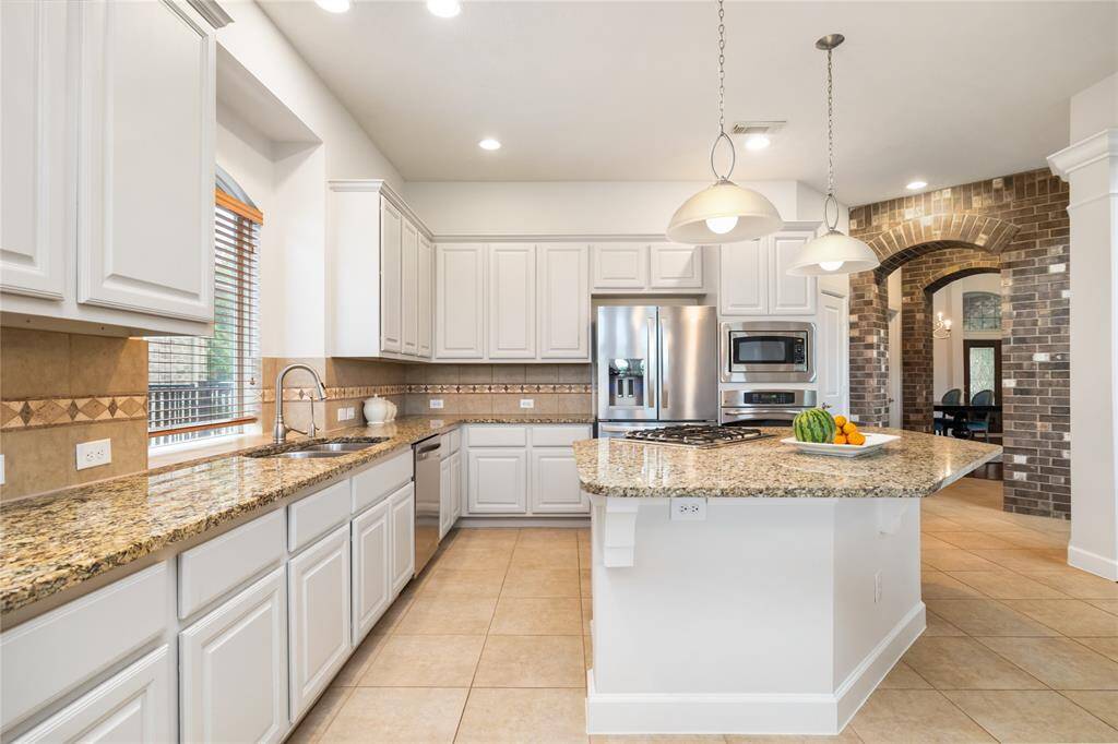 This spacious kitchen features granite countertops, a large island with gas cooktop, stainless steel appliances, and crisp white cabinetry—all with a view into the elegant dining room through a charming brick archway.