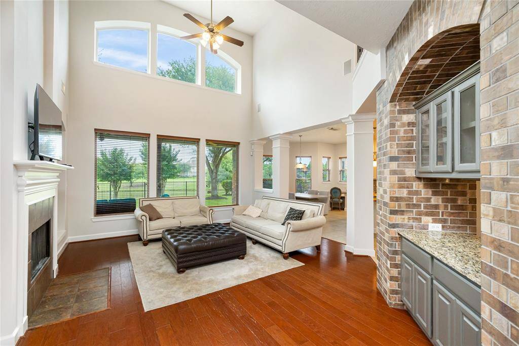 This stunning living area features soaring ceilings, a wall of windows with serene lake views, and rich hardwood floors that flow throughout. A cozy fireplace anchors the space, while the open layout seamlessly connects to the kitchen, breakfast nook, and formal dining room tucked just beyond the brick archway. The spacious primary suite is privately located just off the living room, offering both comfort and convenience in this thoughtfully designed floor plan.