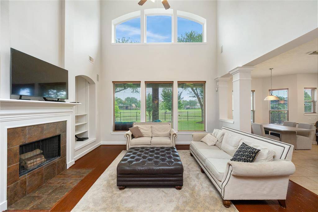 This stunning living area features soaring ceilings, a wall of windows with serene lake views, and rich hardwood floors that flow throughout. A cozy fireplace anchors the space, while the open layout seamlessly connects to the kitchen, breakfast nook, and formal dining room tucked just beyond the brick archway. The spacious primary suite is privately located just off the living room, offering both comfort and convenience in this thoughtfully designed floor plan.