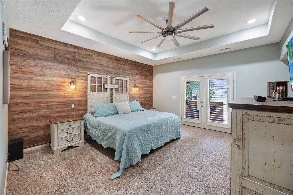The spacious primary bedroom exudes rustic elegance with a feature wall of warm wood look paneling and farmhouse-style sconces flanking the bed. A tray ceiling with recessed LED lighting and a contemporary ceiling fan add height and dimension, while French doors provide private access to the backyard oasis.