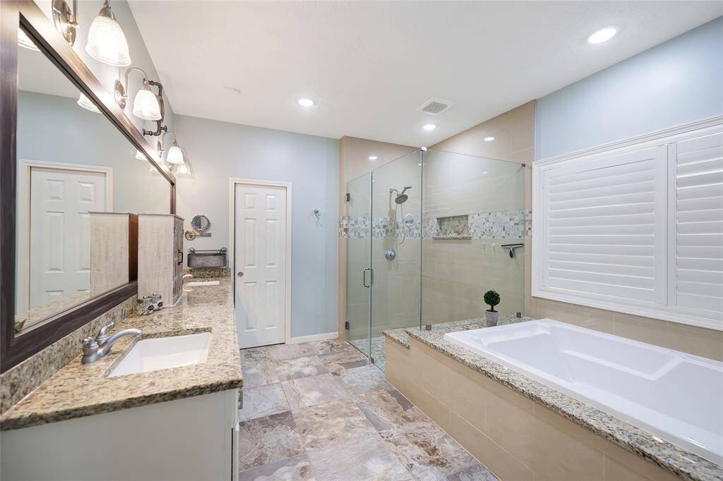 This luxurious bathroom features dual granite-topped vanities, a deep soaking tub with a matching granite surround, and an oversized walk-in shower with frameless glass and a decorative tile inlay. The soft color walls and tile flooring create a relaxing, high-end atmosphere perfect for unwinding.