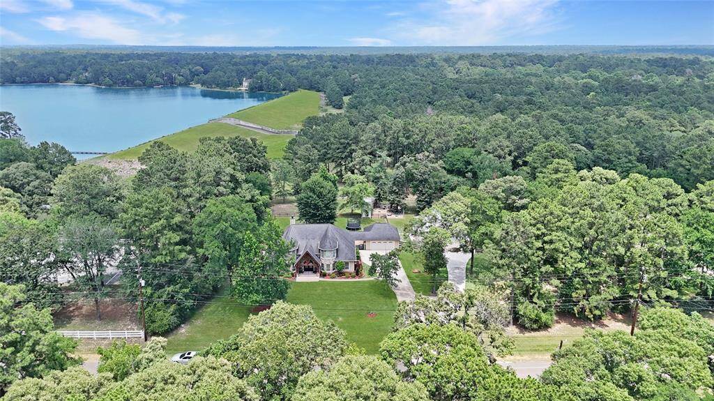 This stunning aerial photo captures the front elevation of the home and surrounding acreage. The home is positioned at the edge of a wooded area, with manicured lawns, a private driveway, and the expansive lakefront just steps away—showcasing a peaceful, picturesque lifestyle.