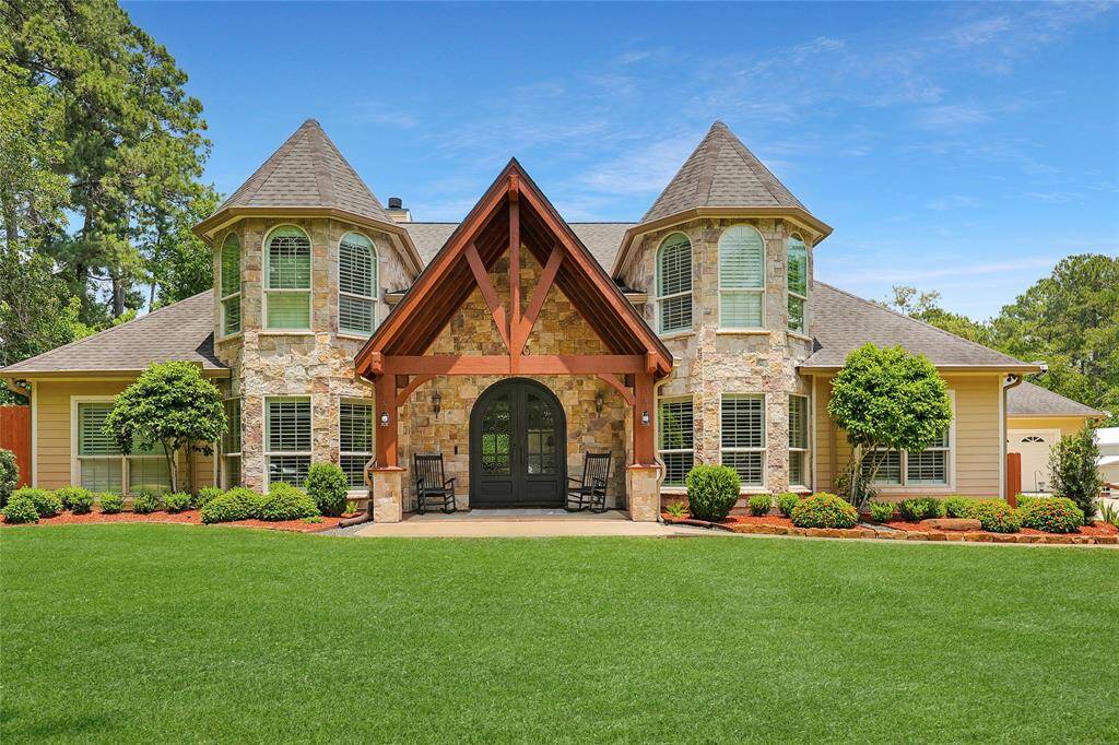 This striking image showcases the front elevation of the home, emphasizing its stone turret architecture and custom wood-beamed portico. The grand arched, iron double doors invite guests in with classic Southern hospitality. Beautiful landscaping with manicured bushes and vibrant mulch beds complete the warm curb appeal.