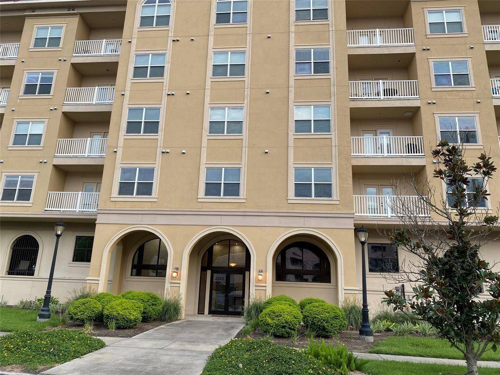 This is a modern multi-story apartment building with a beige exterior and white balcony railings. It features arched entryways and is surrounded by well-maintained landscaping, including shrubs and small trees. The entrance is accentuated by classic lamp posts.