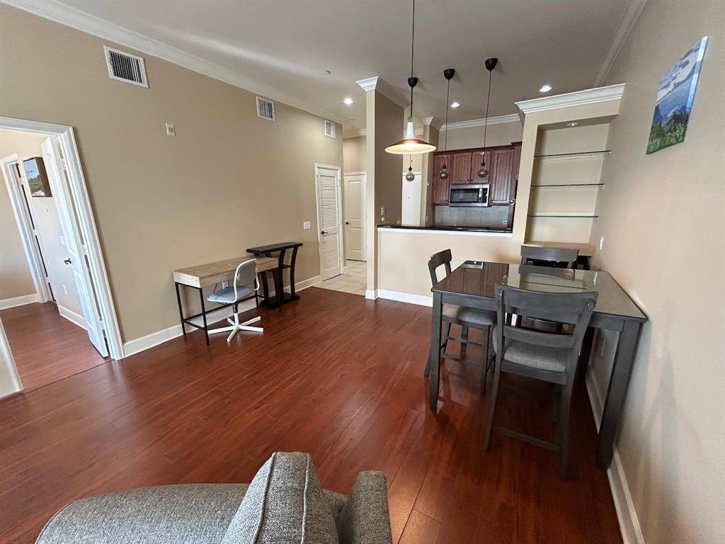 This inviting living space features warm wood flooring, a cozy open plan with a kitchen area, new pendant lighting fixtures, and a breakfast bar. There's a dining table and a small workspace, with neutral walls and ample natural light. Ideal for modern living.