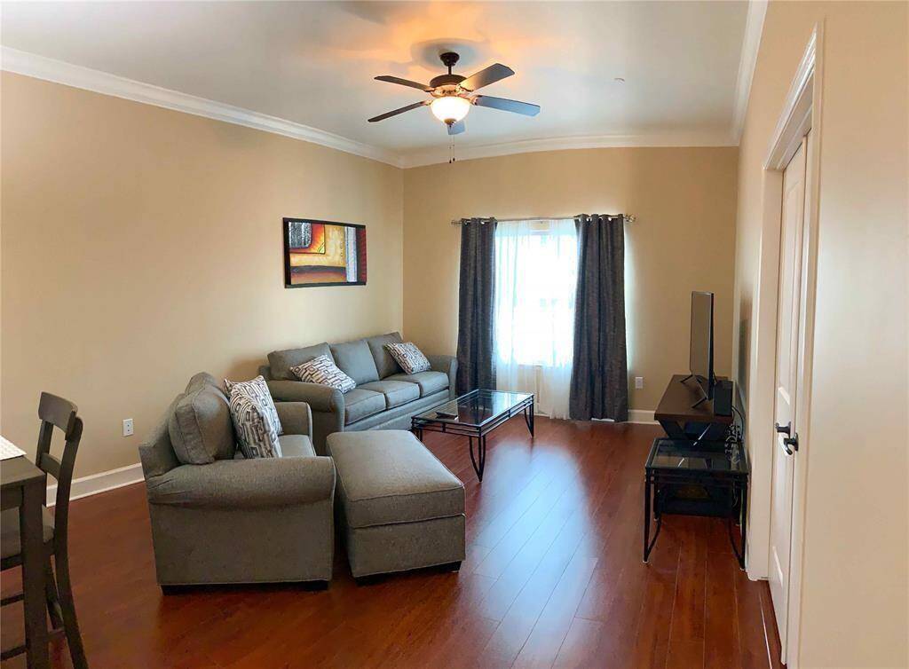 This modern living room with neutral walls, hardwood floors, modern ceiling fan, and ample natural light from a window dressed with dark curtains. The room is furnished with a comfortable grey sofa set and includes a television.