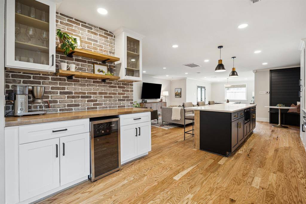This view of the wine and breakfast bar showcases a rich wood countertop paired with a classic brick backsplash. Two floating wood shelves are elegantly framed by glass display cabinets, while below, a built-in wine refrigerator and additional cabinetry.