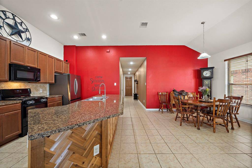 This open-concept kitchen and dining area features sleek granite countertops, warm wood cabinetry, and a bold red accent wall. The space is well-lit with recessed lighting and a large window, creating a bright and inviting atmosphere.