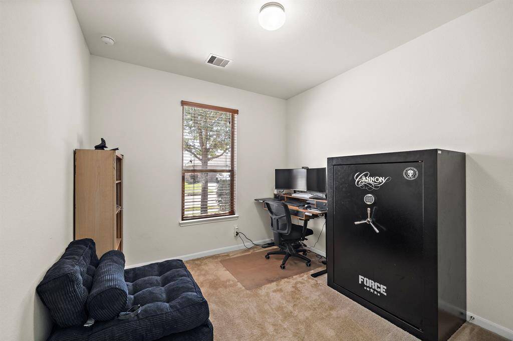 This room is a compact home office with a window providing natural light. It features a large safe, a desk with dual monitors, and a small bookshelf, making it ideal for work and storage.